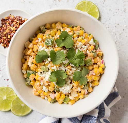Local Street Corn Salad recipe using Vermont sweet corn from Woodstock Farmers' Market. Image shows a white bowl filled with corn, peppers, onions, and cheese, tossed in a light, creamy dressing and topped with sprigs of cilantro. Three lime slices, a small dish of red pepper flakes, and a blue and white striped towel surround the bowl.